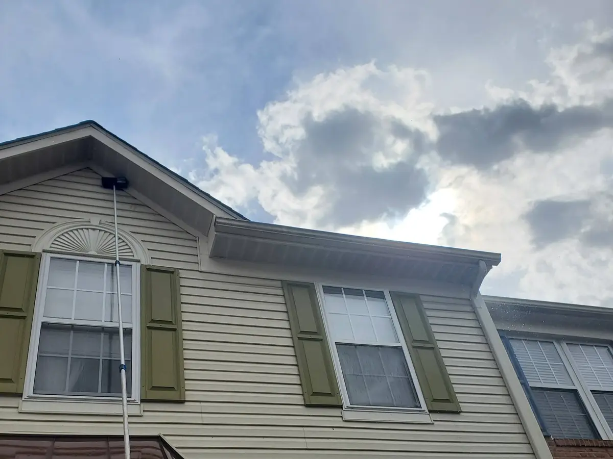 Water-fed pole cleaning the gutter line and upper windows of beige vinyl siding with olive shutters